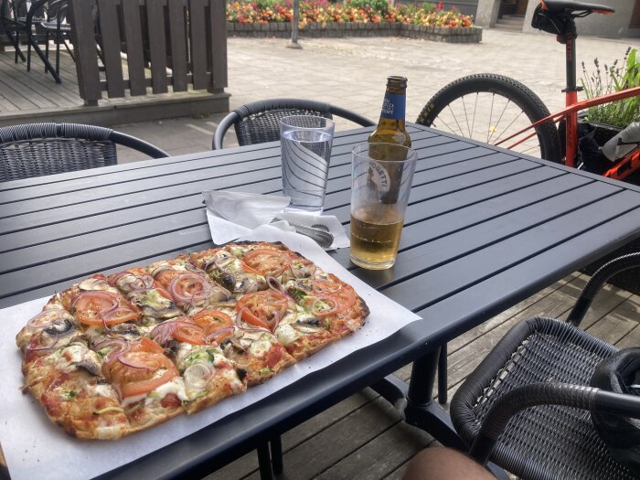 Rektangulär pizza toppad med tomat, lök och svamp på ett svart bord vid en uteservering, med ett glas öl, en flaska öl och ett cykelhjul i bakgrunden.