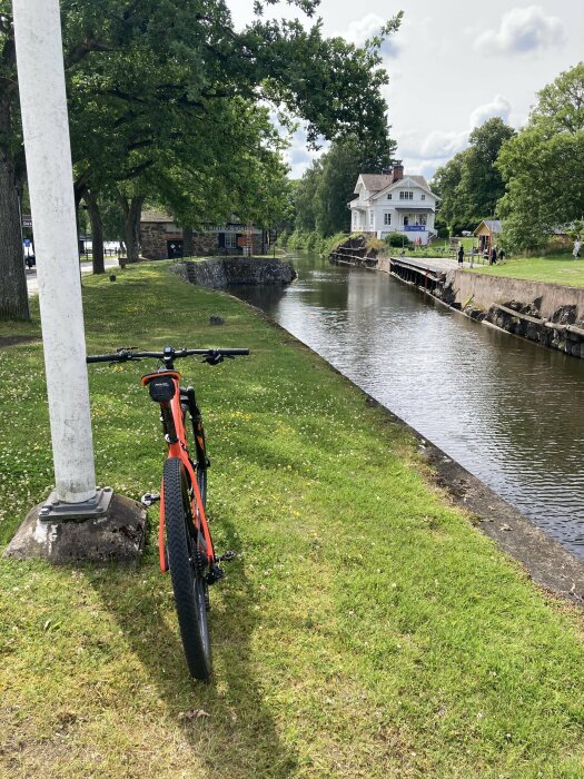 En orange cykel står vid en gräsmatta nära en smal kanal. I bakgrunden finns träd och ett vitt hus längs med kanalen.