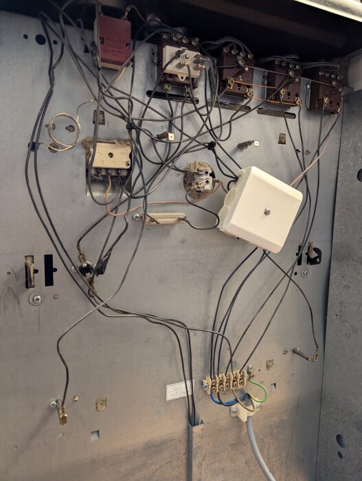 Elektrisk panel med oordnade kablar, varav några är tejpade och ihopbundna med buntband. Panelen har olika komponenter och en vit kopplingsdosa.