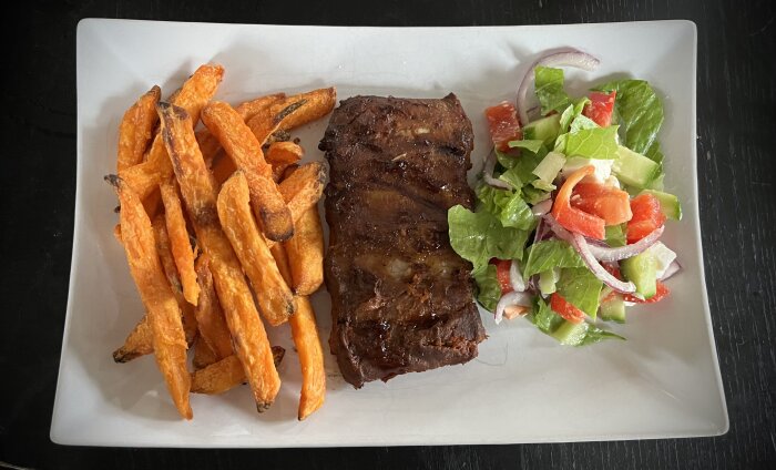 Tallrik med grillade baby back ribs, sötpotatisstrips och sallad med gurka, tomat, rödlök och salladsblad.