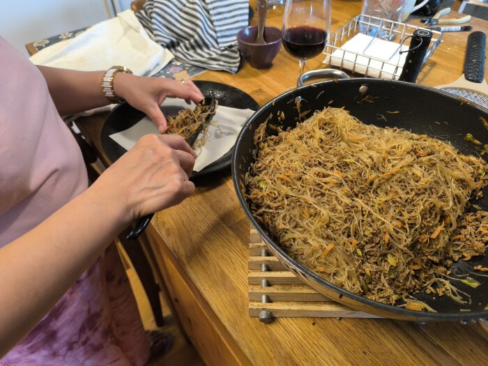 Person rullar vårrullar vid köksbänk, stor panna fylld med nudlar och grönsaker, glas vin i bakgrunden.