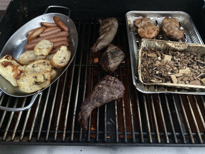 Mat på grill, inklusive korv, grillat bröd, köttbitar, kyckling och träflis i aluminiumform.