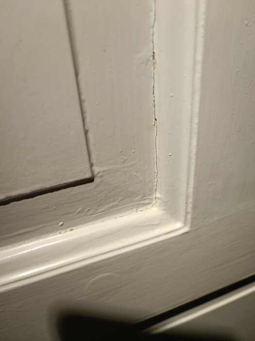 Närbild på en gammal innerdörr med profilerad panel, målad med vit färg som uppvisar sprickor och ojämnheter i ytan.