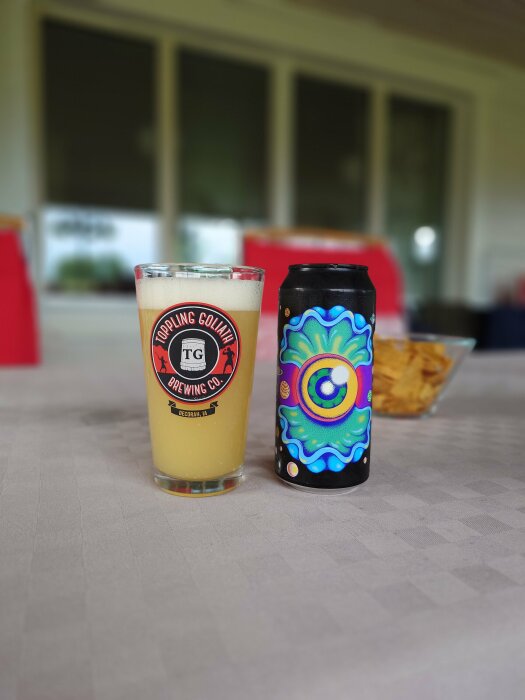 En ölglas med gul vätska märkt "Toppling Goliath Brewing Co." bredvid en färgstarkt designad burk av Omnipollo på ett bord.