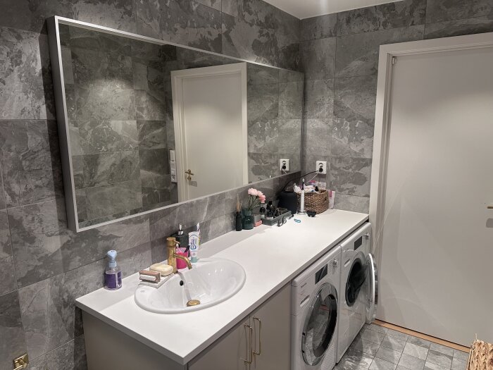 Tvättstuga med stor spegel ovanför en lång bänkskiva med tvättställ; tvättmaskin och torktumlare. Grå väggar, bänken är dekorerad med diverse föremål.