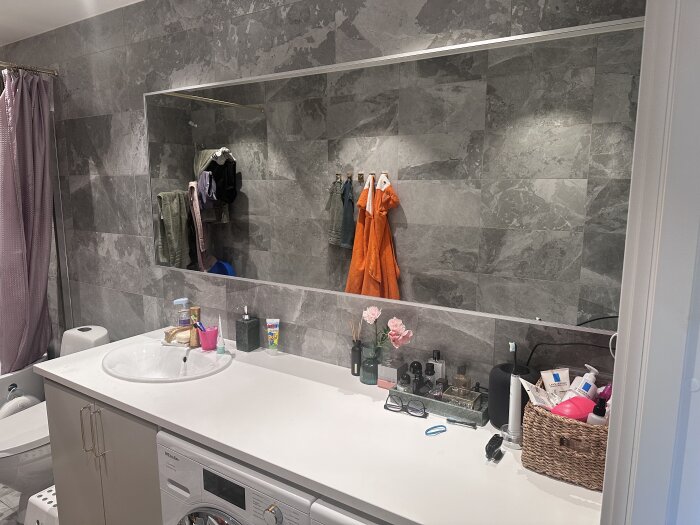 Spegel ovanför en vit bänkskiva med handfat, omgiven av gråa stenväggar i ett badrum. Bänken har toalettartiklar, blommor och andra småföremål.