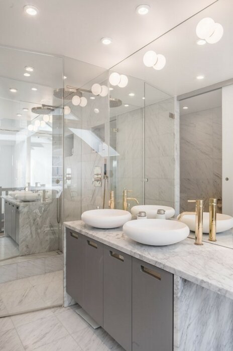 Modernt badrum med grå skåp, en stor spegelvägg, två vita, rundade handfat på en marmorskiva och flera takbelysningar med vita glasklot.