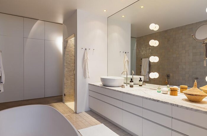Modernt badrum med stor spegel ovanför en lång vit marmorbänk med tvättställ, klotbelysning på väggen och flera badrumsartiklar på bänken.