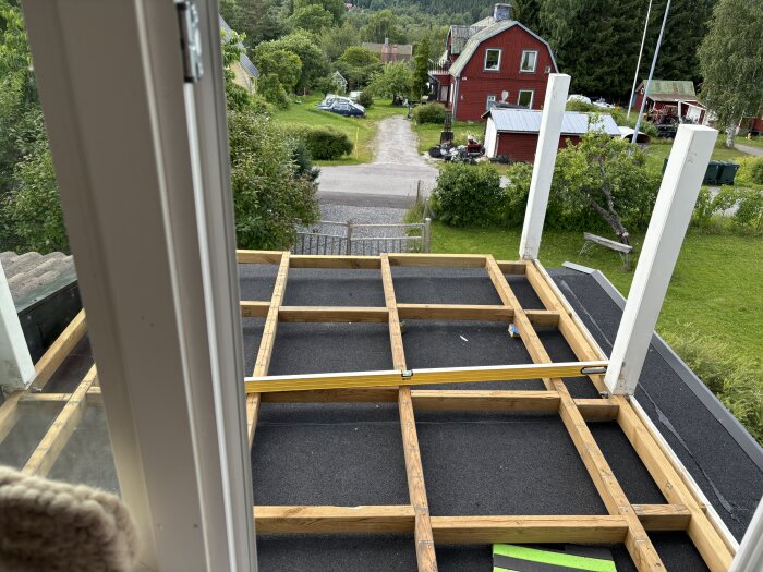 Pågående byggprojekt av balkong fotograferad från ett fönster, med exponerade träramar och två uppställda pelare, omgivande grönska och röda hus i bakgrunden.