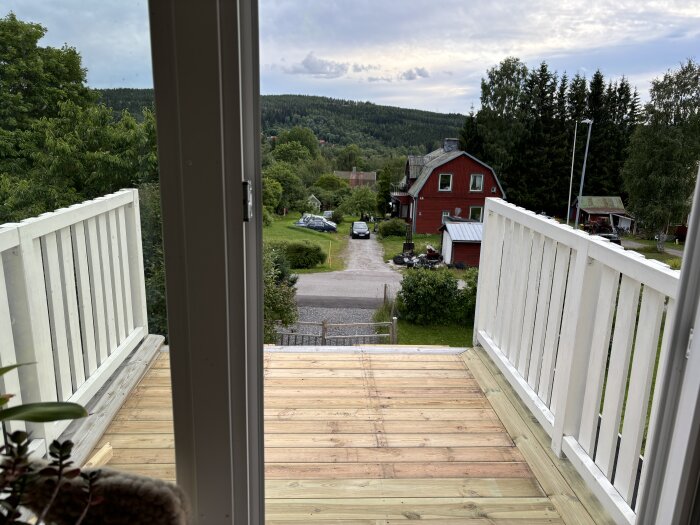 Halvfärdig balkong med trägolv och vitmålade räcken, sett genom en glasdörr. Landsbygd med röda hus och skog i bakgrunden.