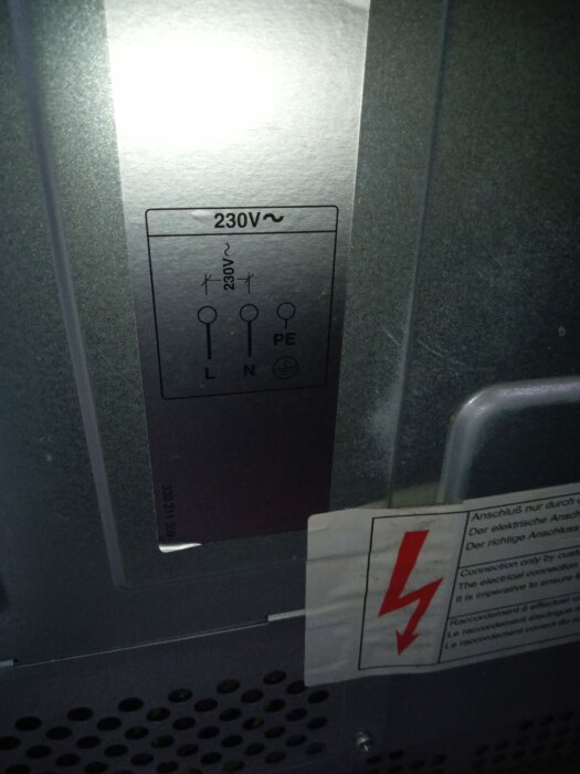 Baksidan av en ugn med märkningen "230V" och symboler för anslutningarna L, N och PE, samt en varningsetikett för elektrisk fara.