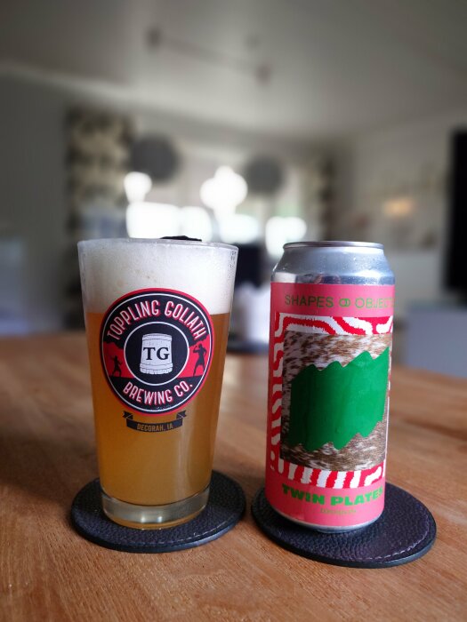 Ett glas öl från Toppling Goliath Brewing Co. bredvid en burk Twin Plates Ddh Hazy Ipa på ett träbord, inomhus med suddig bakgrund.