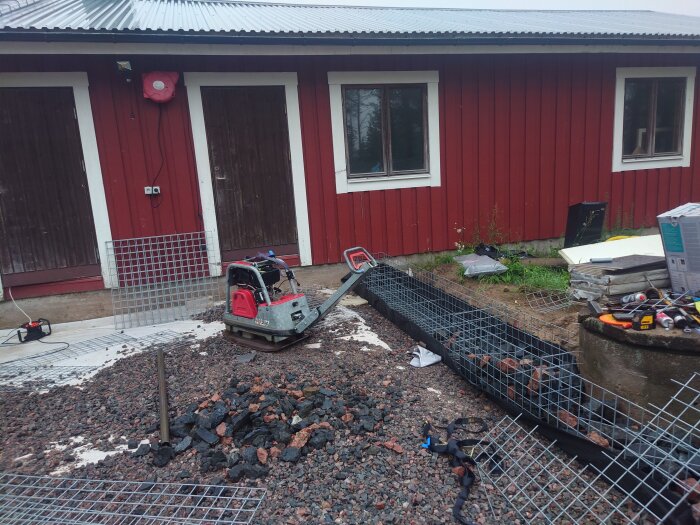 Markarbetsprojekt framför ett rött hus med staplade gabioner, en markvibrator, och spridda byggmaterial.