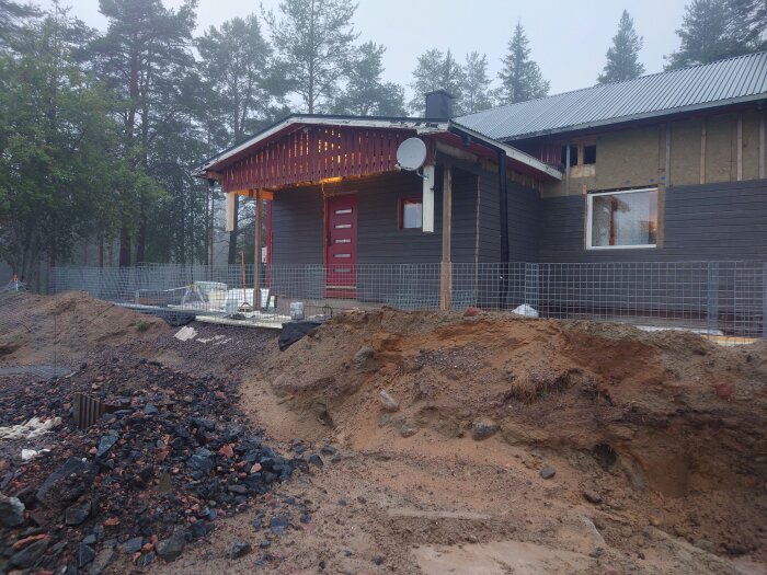 En renoverad byggnad med ny ytterpanel, en röd dörr och ett stort grävt område framför. Gräsmatta och skog i bakgrunden. Byggmaterial synligt runt huset.