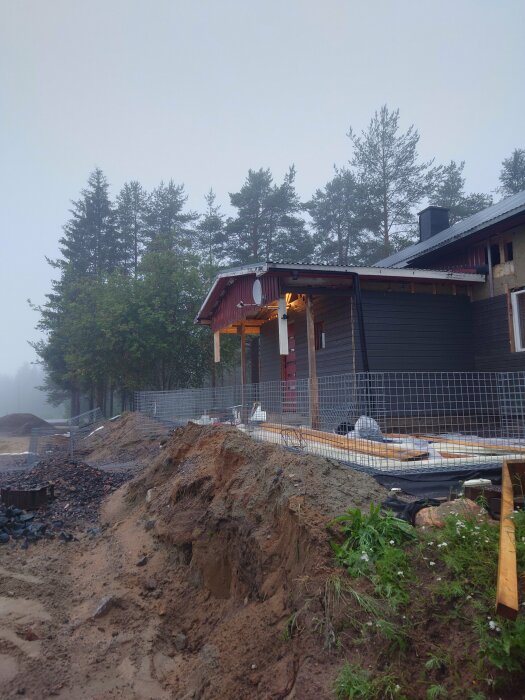 Byggarbetsplats med ett hus, staket av gabionkorgar, och uppgrävd mark framför träd i bakgrunden. Verktyg och material ligger på marken.