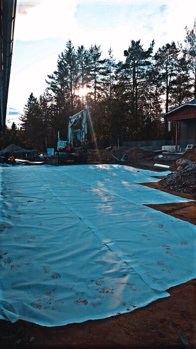 Grävmaskin på stor inhägnad gårdsbeklädnad med geotextilduk, omgiven av träd och ett garage. Ordentligt markförberedande arbete pågår under solnedgången.