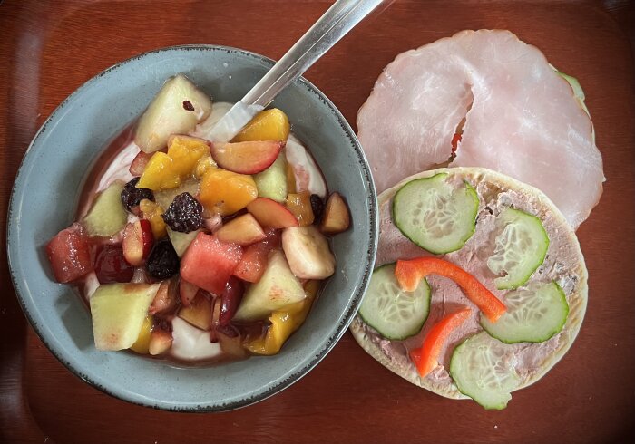 Skål med kvarg och fruktsallad innehållande björnbär, plommon, meloner, nektarin, banan och mango bredvid tunnbrödsrulle med gurka, paprikastrimlor och skinka.