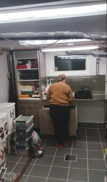 Kvinna i brun tröja står vid köksbänken i ett hundrum med förvaringshyllor och ett genomfärgat klinkergolv.