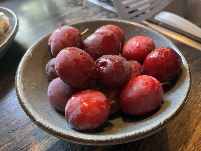 En grå skål fylld med mörkröda plommon från ett träd, placerad på ett mörkt träbord.