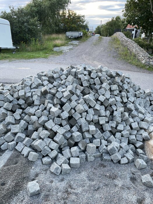 En stor hög med smågatsten som ska användas för att lägga stenläggning på en uppfart. I bakgrunden syns en grusväg, grönska och en mur.