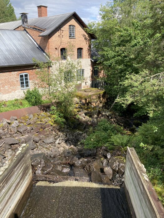 Tegelhus med plåttak nära ett vattendrag omgivet av stenblock och grönska, sedd från ovanför en träkonstruktion med rinnande vatten.