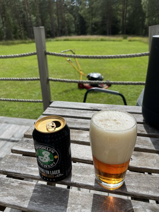 En ölburk och ett ölglas med Brooklyn Lager på ett träbord utomhus, med en gräsklippare stående på en gräsmatta i bakgrunden.