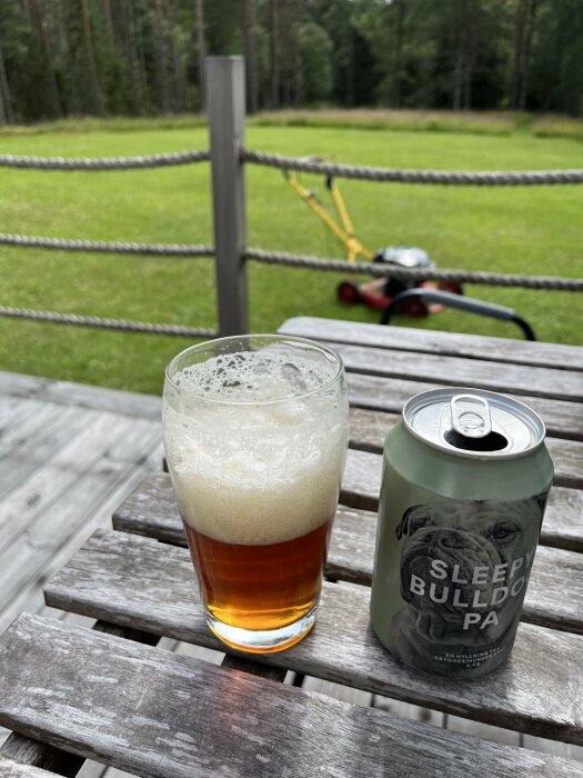 Ett glas öl och en ölburk på ett träbord, med en gräsklippare och en grön gräsmatta i bakgrunden.