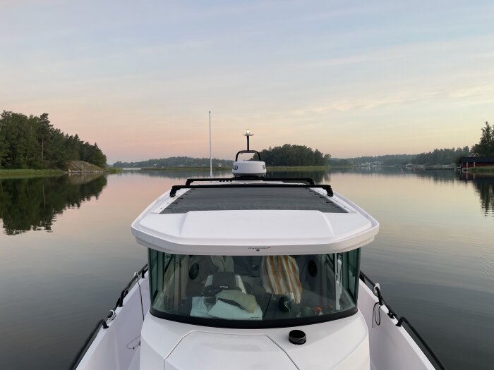 En båt åker genom en lugn sjö eller hav vid solnedgång eller soluppgång med skogar och hus längs stranden.