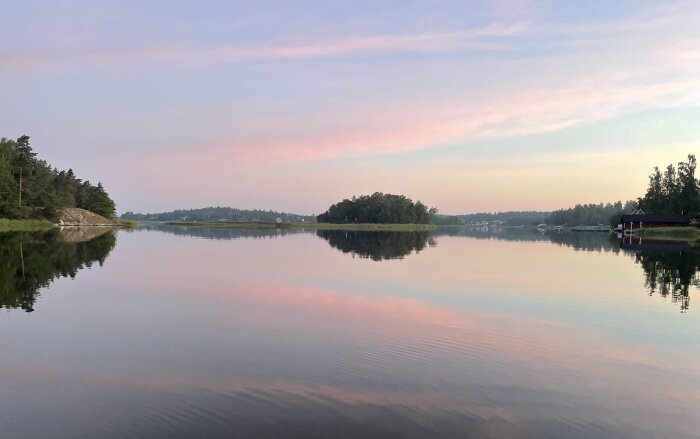 Sjölandskap en tidig morgon med spegelblankt vatten och rosa, lila och blå nyanser på himlen, omgiven av skog och bryggor.