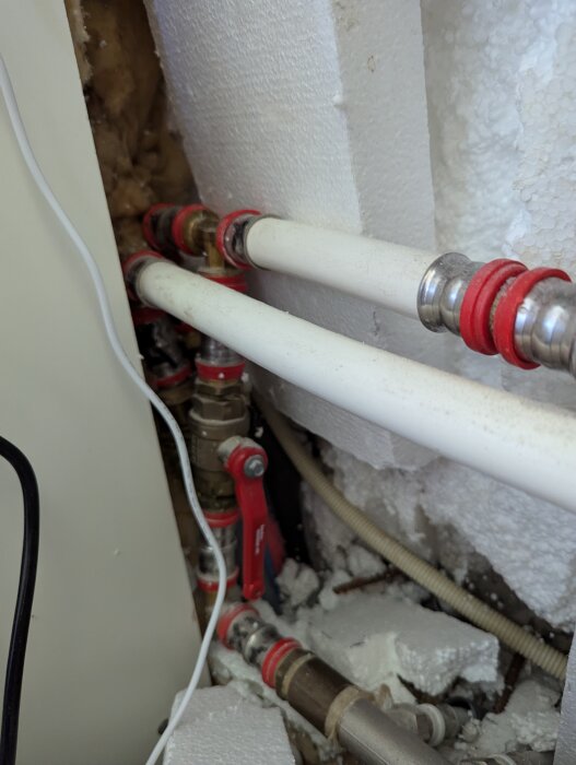Två vita rör med röda kopplingar vid ett skåp fyllt med frigolit, bilden visar även en röd ventil och några elektriska kablar.