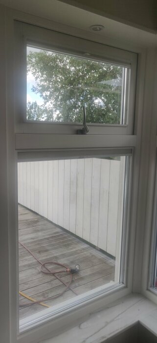 Fönster med landskapsvy som visar träd, staket och en terrass med en vattenslang liggande på golvet. Frågande om justering av fönster underkant.