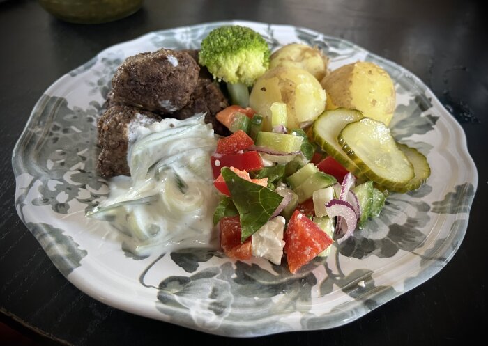 Tallrik med köttbullar toppade med tsatsiki, inlagd gurka, kokt potatis, broccoli och fetaostsallad med paprika, gurka, rödlök, tomat och salladsblad.