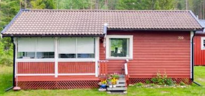 Rött fritidshus med inglasad altan och blomkrukor vid ingången, beläget i en grön trädgård med träd i bakgrunden. Husets tak är täckt med bruna takpannor.