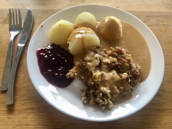 Tallrik med kålpudding, tre potatisar, brun sås och lingonsylt, fotad bredvid bestick på träbord.