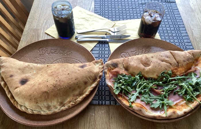 Två stora inbakade pizzor på tallrikar med glas med cola och is, samt bestick på gula servetter på ett matbord med grå bordstablett.