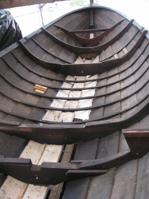 Träbåt under byggnation med synliga spant och bordläggning, där några träplankor och verktyg delvis täcker durken.