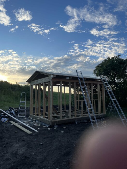 En träbyggnad under uppbyggnad med råspont på taket, stegar lutade mot stommen och byggmaterial på marken under en blå himmel med moln.