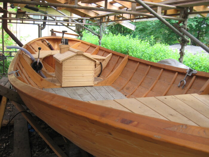 Träbåt under renovering med synliga verktyg och reglar i en träkonstruktion, omgiven av grönska och delvis täckt takstruktur.