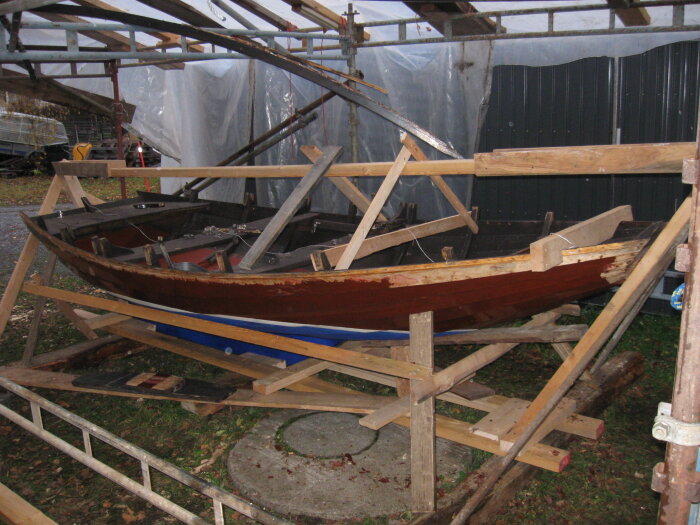 Träbåt under reparation med stödstrukturer runt omkring, troligen en klinkbyggd båt av inhemskt virke. Plats: en skyddad utomhusarbetsplats.