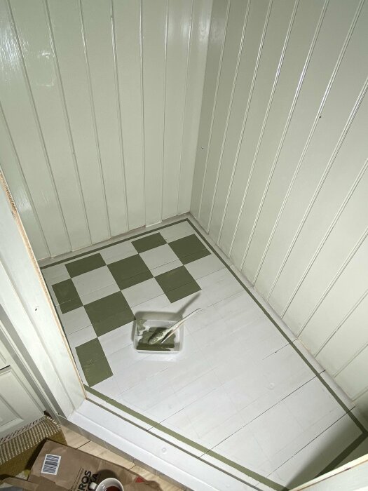 Målat schackrutigt golv i skafferi, vita och gröna rutor. Målarfärg och pensel i en vit bricka ligger på golvet. Panellister på väggarna.