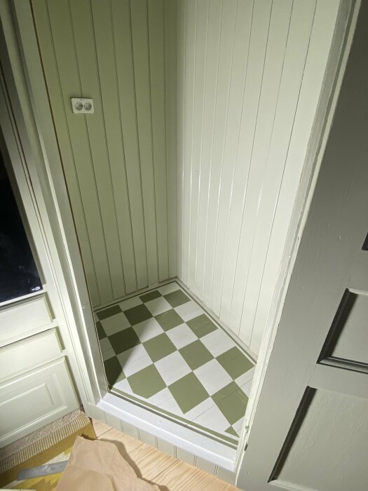 Nymålat skafferigolvet med gröna och vita schackrutor i ett kök. Väggar och skafferidörr i ljus träpanel.
