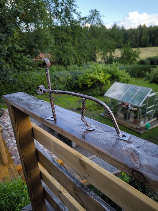 Handgjord krok av armeringsjärn monterad på ett staket, lantlig bakgrund med grönska och växthus.