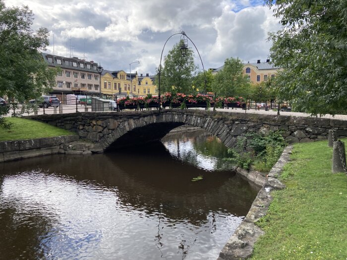 En stenbro över en lugn vattenkanal med byggnader och blomsterarrangemang i bakgrunden, under en molnig himmel.