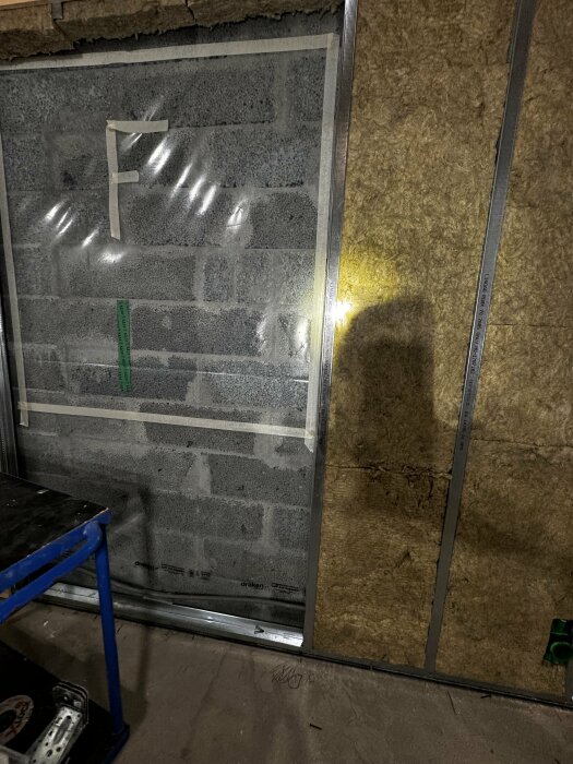 Isolerad källarvägg med lecablock, plastfilm som ångspärr, stålskenor och delvis monterad mineralull. En ljuskälla lyser på väggen till höger.