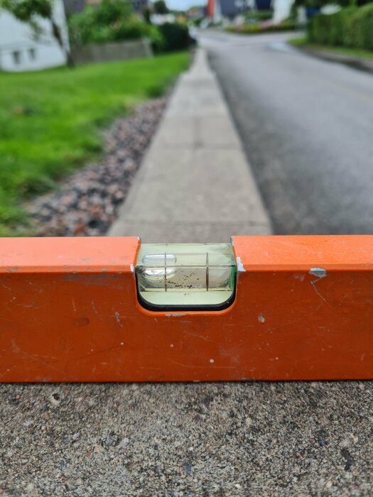 Orange vattenpass på mur, bubblan indikerar en lutning utåt. Bakgrunden visar en trottoar och grönska vid en gata med hus.