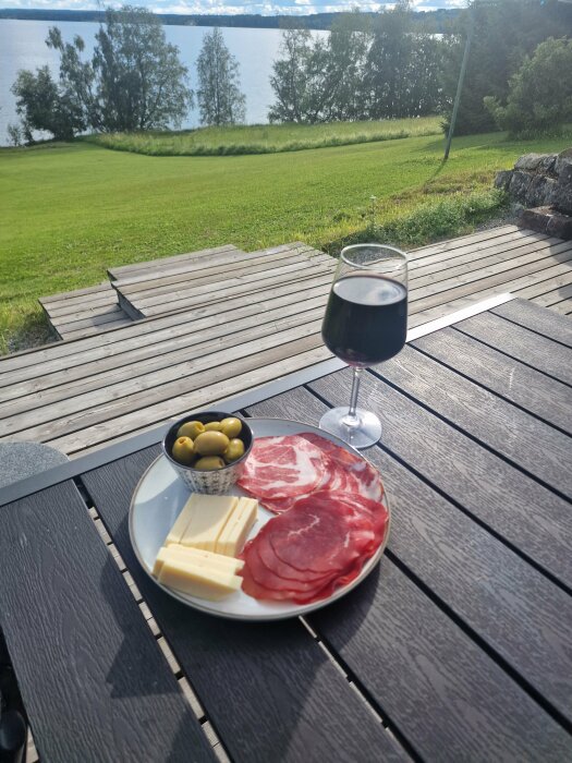 Ett fat med ost, charkuterier och oliver samt ett glas rödvin på ett bord med utsikt över en gräsmatta och sjö i bakgrunden.