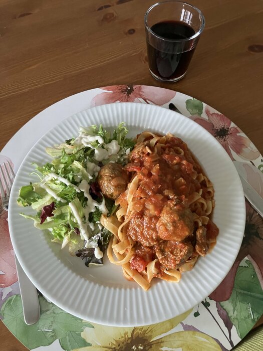 En tallrik med italienska köttbullar i tomatsås, pasta och en grönsallad med vitlökssås. Ett glas röd dryck bredvid tallriken på ett blommigt underlägg.