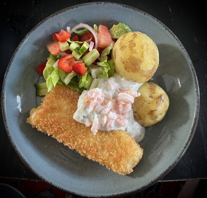 Tallrik med panerad torskfilé, citronsås med dill och räkor, kokt färskpotatis samt en sallad med blandade grönsaker.