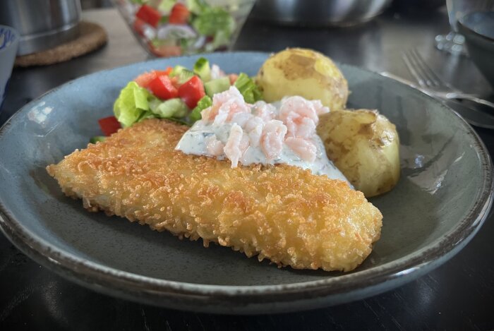 Panerad torsk med citron- och dillsås, kokt färskpotatis, sallad och räkor på toppen, serverad på en tallrik.