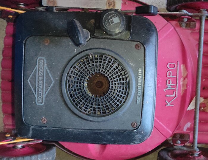 Rosa gräsklippare med en svart motor, märkt "Briggs & Stratton", och text "KLIPPO" på klipparhöljet. Motorlocket har ventilationsgaller och tanklock.